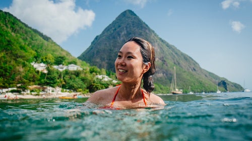 A woman swims in the Caribbean waters of Saint Lucia, with one of the green Piton mountains in the background.