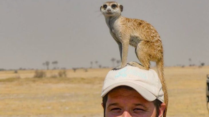 A meerkat sits atop a man wearing a ball cap and scans the horizon of Botswana’s Makgadikgadi Pans.