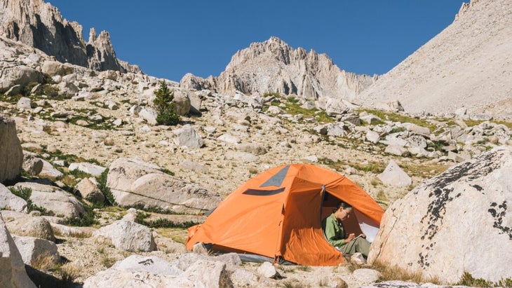 A woman sits in her orange tent, writing in a journal. She's pitched her tent on a hillside with jagged peaks soaring above.