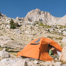 A woman sits in her orange tent, writing in a journal. She's pitched her tent on a hillside with jagged peaks soaring above.