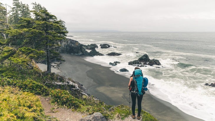 A woman carrying a big backpack looks over the black-sand shoreline and wind-blown trees of British Columbia’s West Coast Trail.