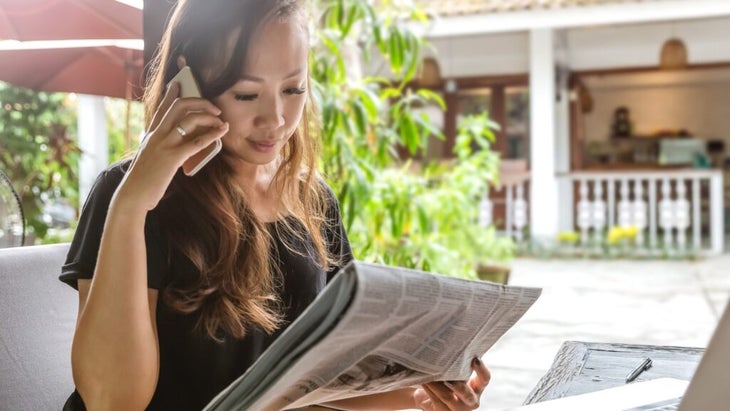 A woman scans the newspaper classifieds while making a call on her cell phone. 