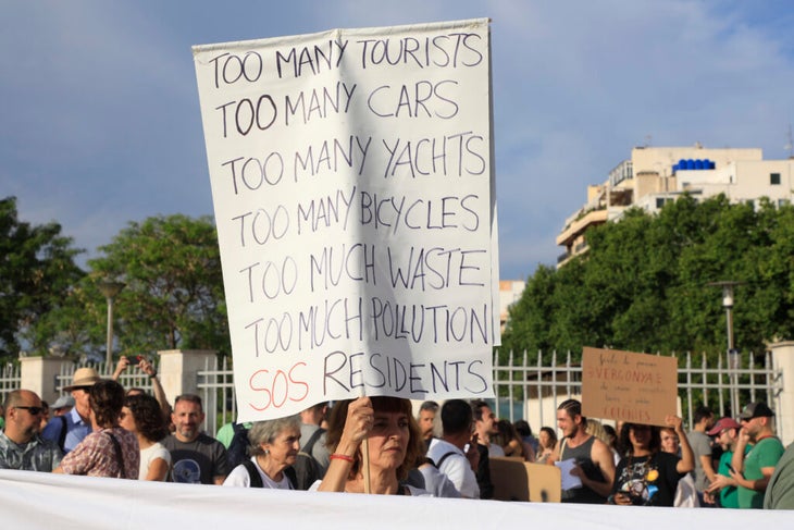 A local of Palma, Spain, holds up a sign that reads "Too many tourists. Too many cars. Too many yachts. Too many bicycles. Too much waste. Too much pollution. SOS Residents."