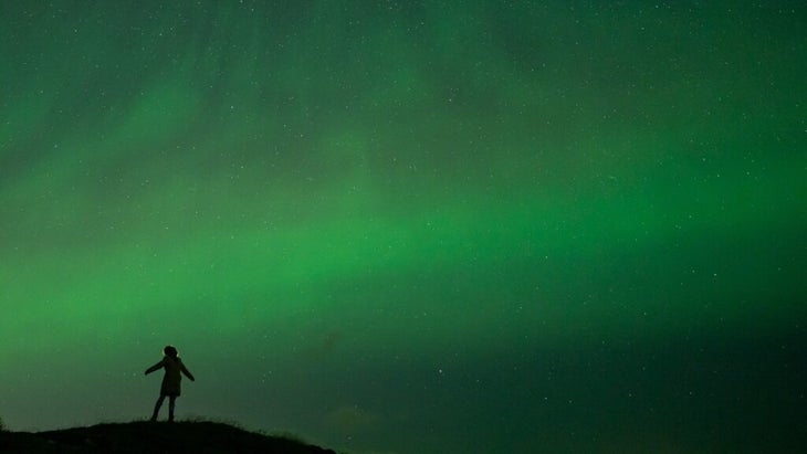 A silhouette of the author on a hillside in Iceland, with a green ribbon of northern lights and a sparkling sky behind her. She took the photo using a timer.