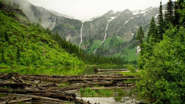 Thin waterfalls drop down into Avalanche Lake, which is surrounded by green hills and brush. The lake is just five miles east of Glacier National Park's Lake Macdonald Lodge.