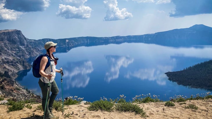 The author in hiking attire and carrying poles, looking over a glassy blue Crater Lake. 