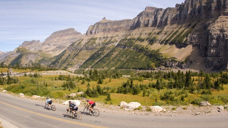 Three road cyclists whiz down a traffic-free Going-to-the-Sun Road in Glacier National Park, Montana, with towering mountains to their right.