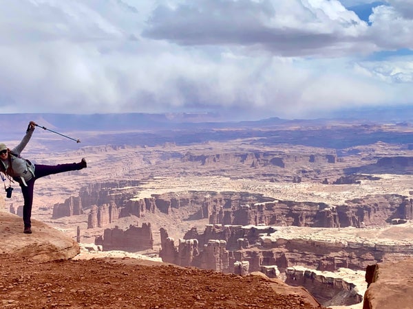 The author holding hiking poles jokes like she's about to cartwheel into the huge canyon below Grand View Point in Canyonlands National Park.