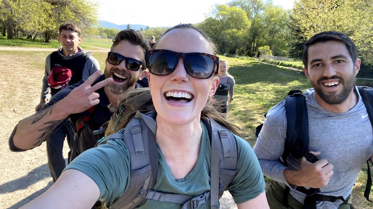 A woman takes a selfie in a green park with three men. They are all wearing backpacks.