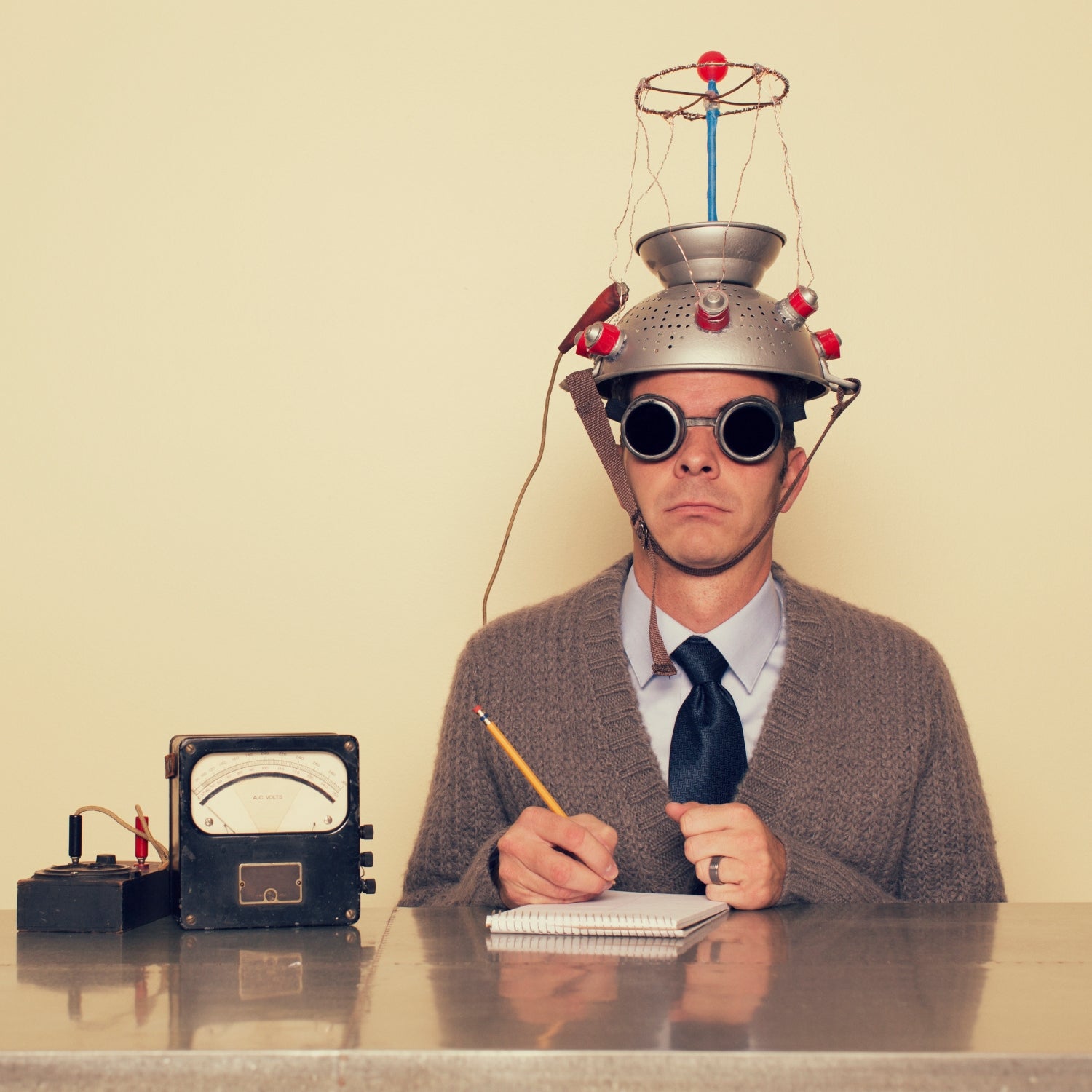 A scientist wears a metallic brain wave contraption on his head and sits behind a desk taking notes