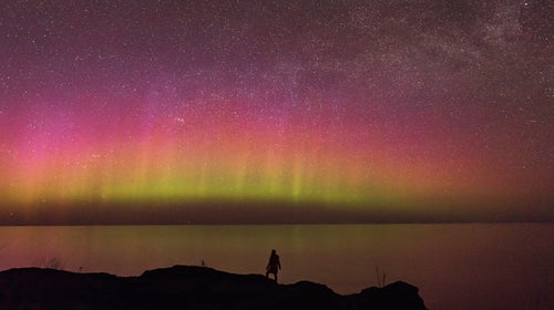 A silhouette of the author standing on a hillside of Wisconsin’s Keweenaw Peninsula looking over a still Lake Superior, with a gorgeous aurora of purple, pink, yellow and green on the horizon.