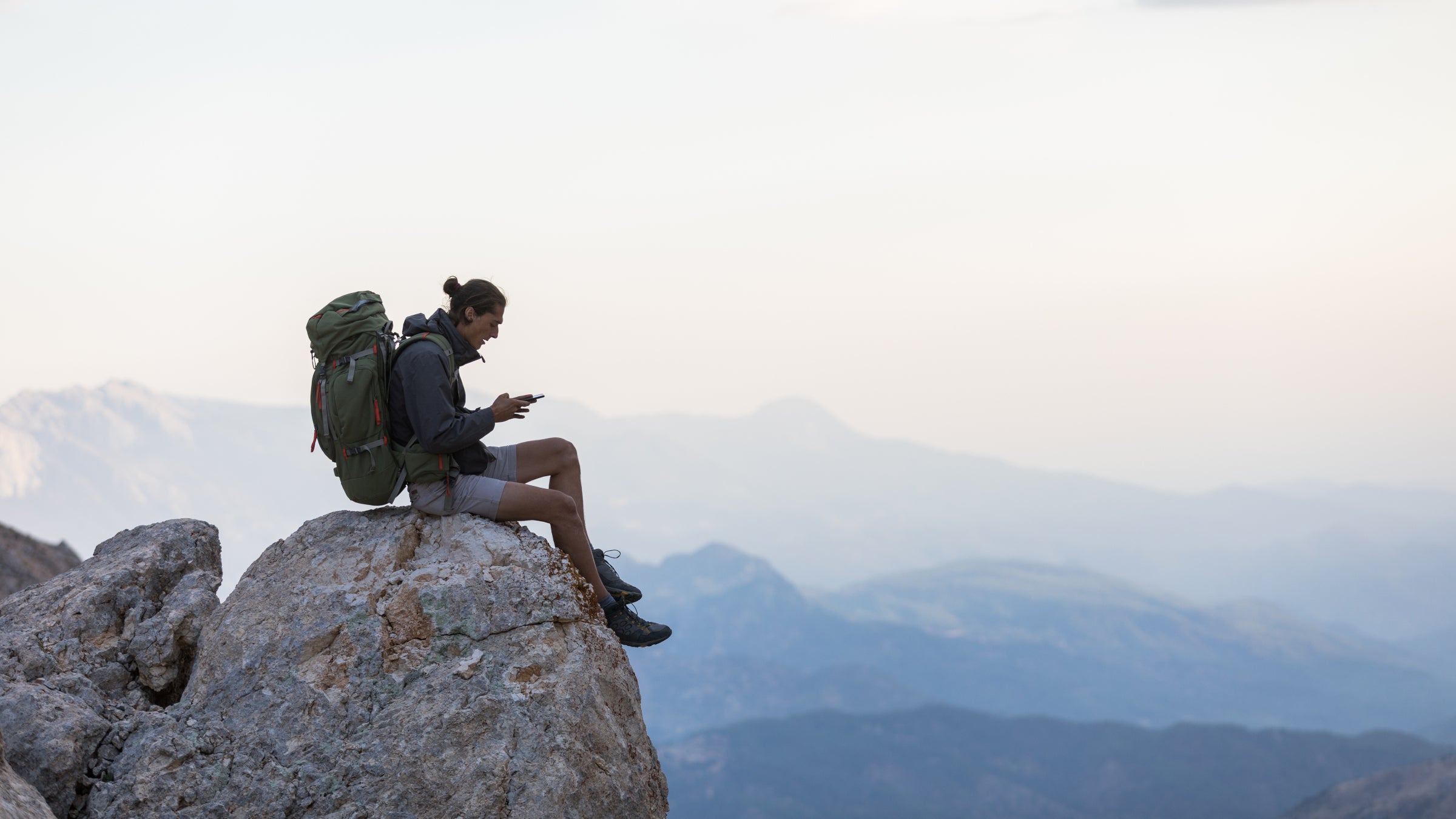 A hiker sitting on a rock looking at his phone with mountains in the background