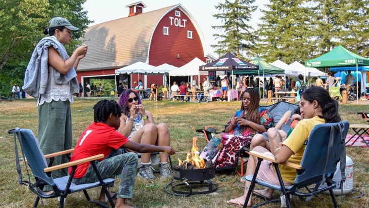 A group of people gather around an outdoor fire pit, eating and enjoying a sunny day at the Refuge Outdoor Festival in Washington. Behind them is a red barn and several tents set up with vendors and a crowd.