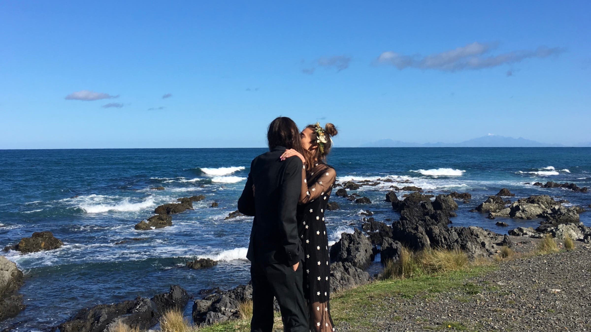 A man and woman in formal clothing embrace while standing next to a rocky shore