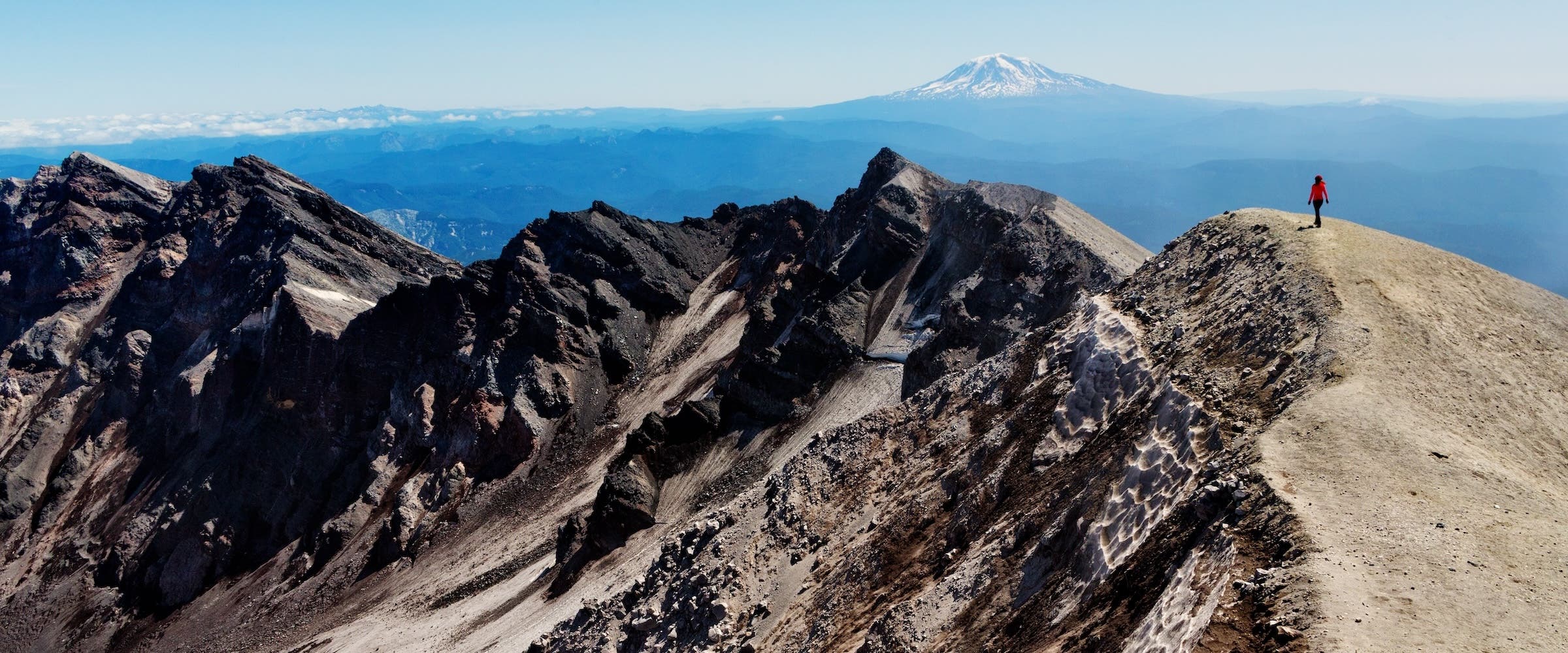 Back view of hiker walking along edge of volcanic crater at Mount St Helens summit with Mount Adams on horizon in distance, summer afternoon, Washington state