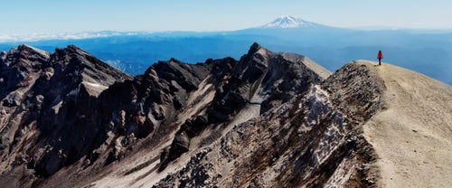 Back view of hiker walking along edge of volcanic crater at Mount St Helens summit with Mount Adams on horizon in distance, summer afternoon, Washington state