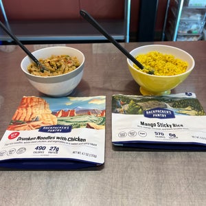 Two freeze-dried backpacking meal packets next to bowls of reheated dinners.