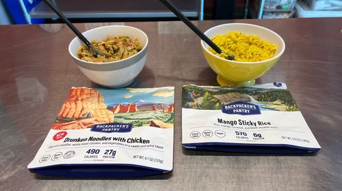 Two freeze-dried backpacking meal packets next to bowls of reheated dinners.