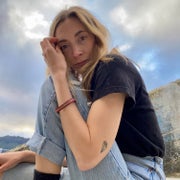 A blonde woman sits outside wearing a black t-shirt and cuffed blue jeans.