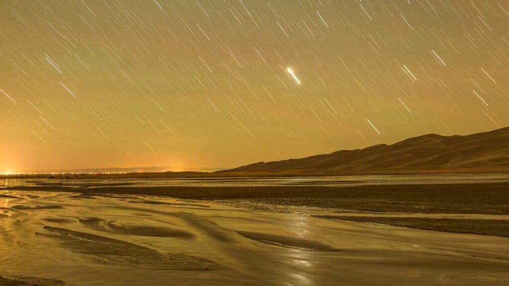 A glowing night sky with a shooting star over Colorado’s Great Sand Dunes National Park and Medano Creek