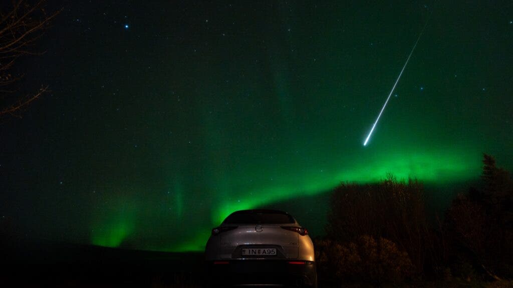 A car parked on the road on a dark night in Iceland, with the northern lights glowing green on the horizon and a fireball shooting down from the sky.