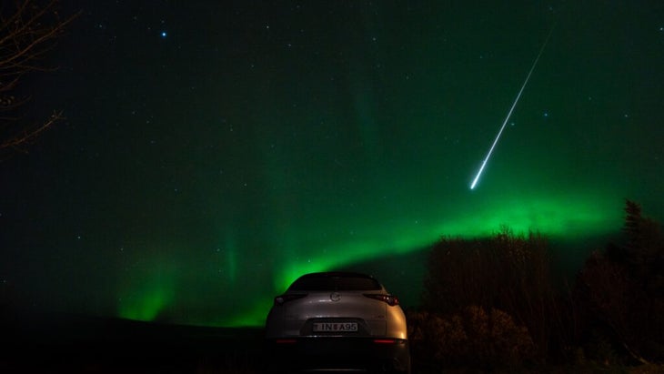 A car parked on the road on a dark night in Iceland, with the northern lights glowing green on the horizon and a fireball shooting down from the sky. 