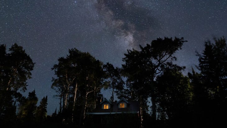 One of Michigan’s Fresh Coast Cabins at night, surrounded by a few trees, with the Milky Way stretching across the sky above.