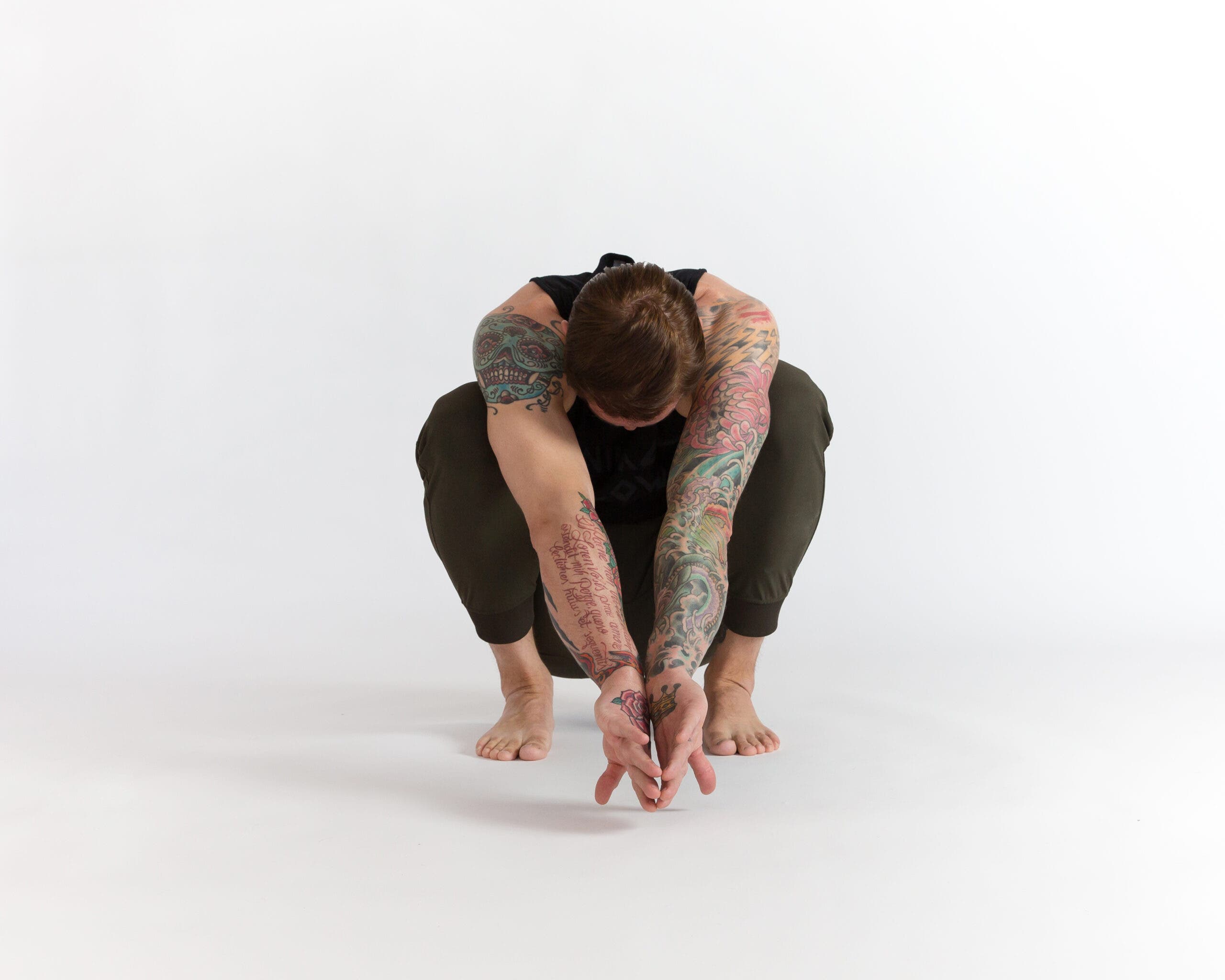 Man in ape reach. He is in a deep squat with his arms turned inward and outstretched in front of him, backs of the hands touching. His gaze is downward.