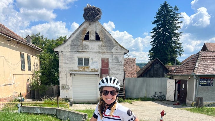 The author stops her bike ride in Romania to pose in front of a house where a muster of storks has constructed a large nest atop a building.