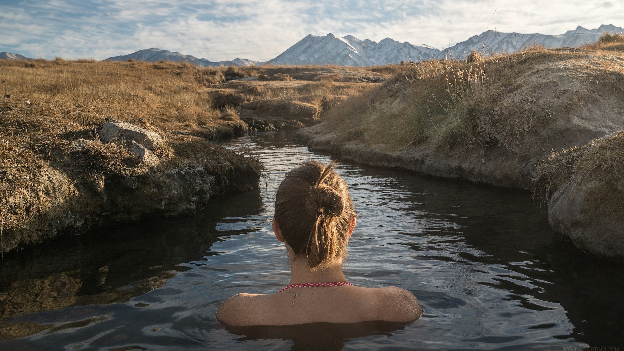 woman soaks in a warm stream and gazes at a mountain view