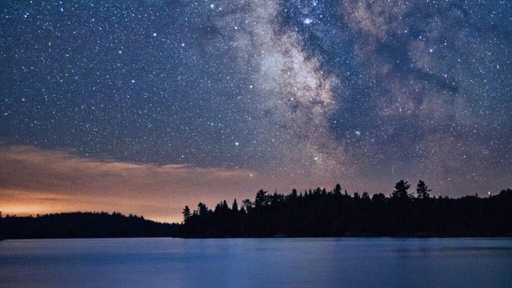 The Milky way shines bright over a silhouetted skyline of a low-lying island of trees and a lake at Voyageurs National Park.