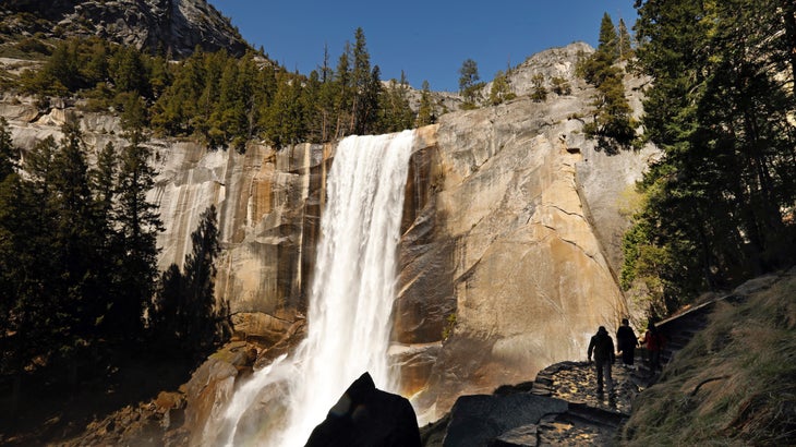 A pair of hikers head up trail steps, with a raging Vernal Fall pours off the granite cliffs at Yosemite National Park.