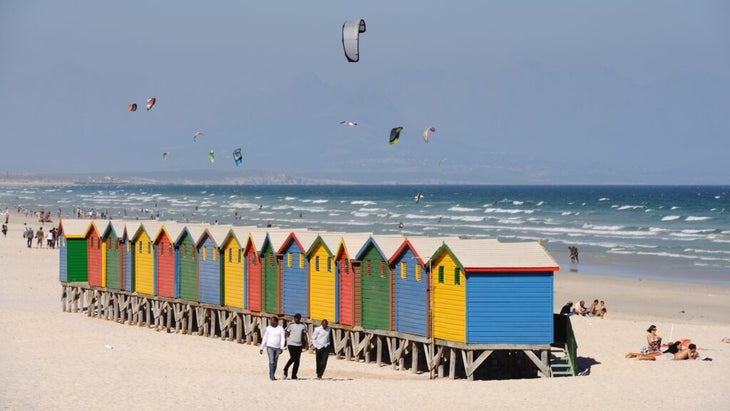Colorful changing houses line South Africa’s Muizenberg Beach, near Cape Town, while kitesurfers play in the surf.