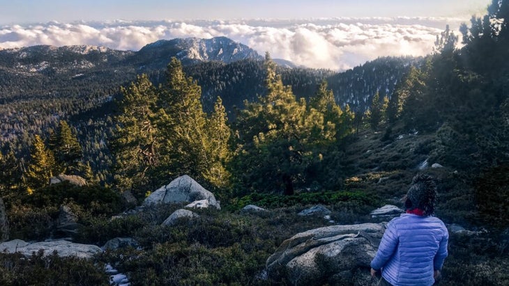 The writer, wearing a jacket and beanie, looks over a panorama of Southern California's forested horizon, covered by clouds at sunset.