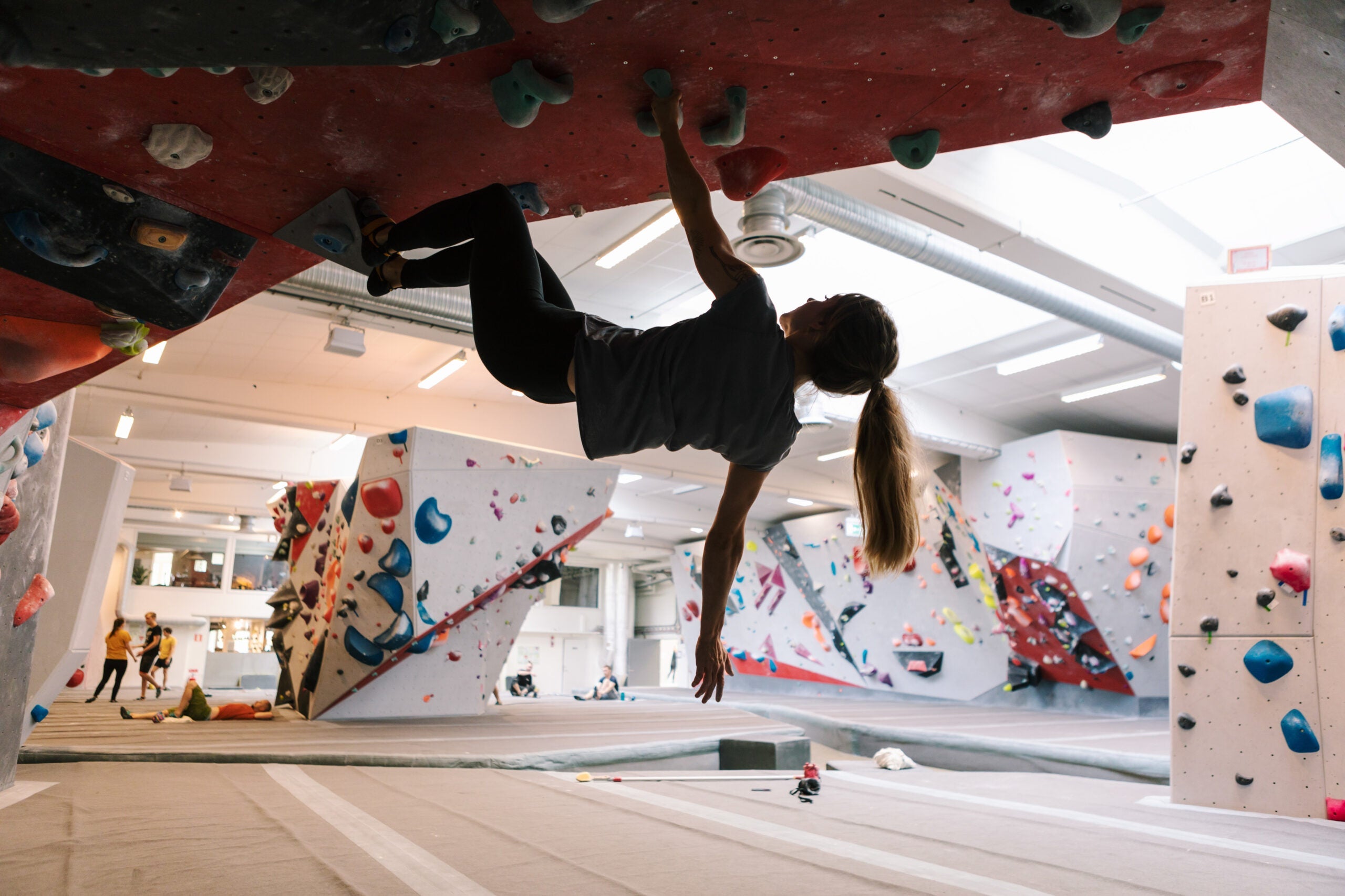 A woman climbing an overhanging in a semi-crowded rock climbing gym.