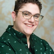 A portrait of Julie Kliegman. They have short brown hair and glasses, and are smiling in front of a floral background. They are wearing a patterned green shirt with a collar.