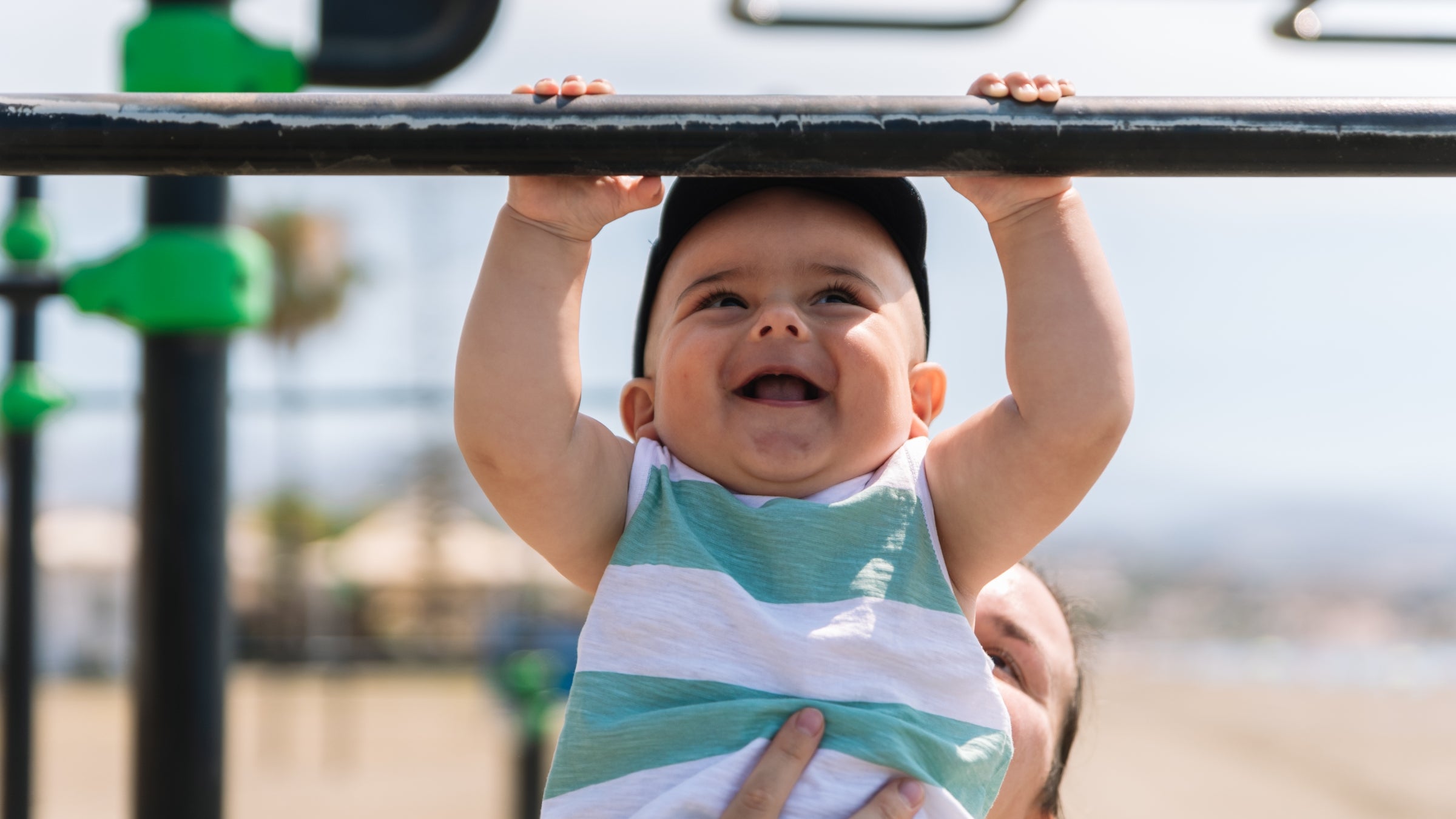 A baby holds onto a pull-up bar while being held by his mother.