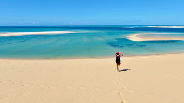 A woman has walked down the soft golden sand to the Atlantic waterfront of one of Mozambique's Bazaruto archipelago. The water is swirled various colors of blues and shows two nearby white sandy islets.