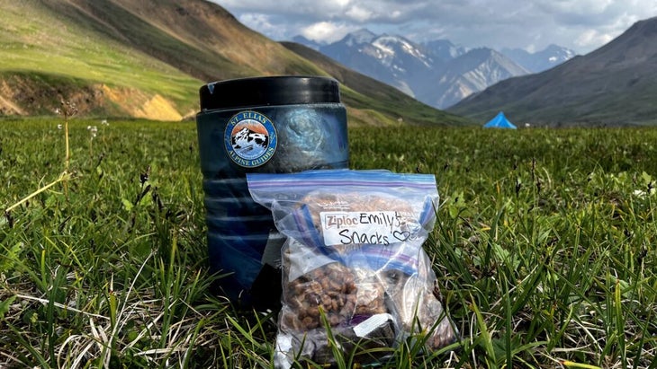 A filled bear can and a ziplock bag, both filled with snacks, set on a field in the Alaskan backcountry, with a tent on the horizon. 