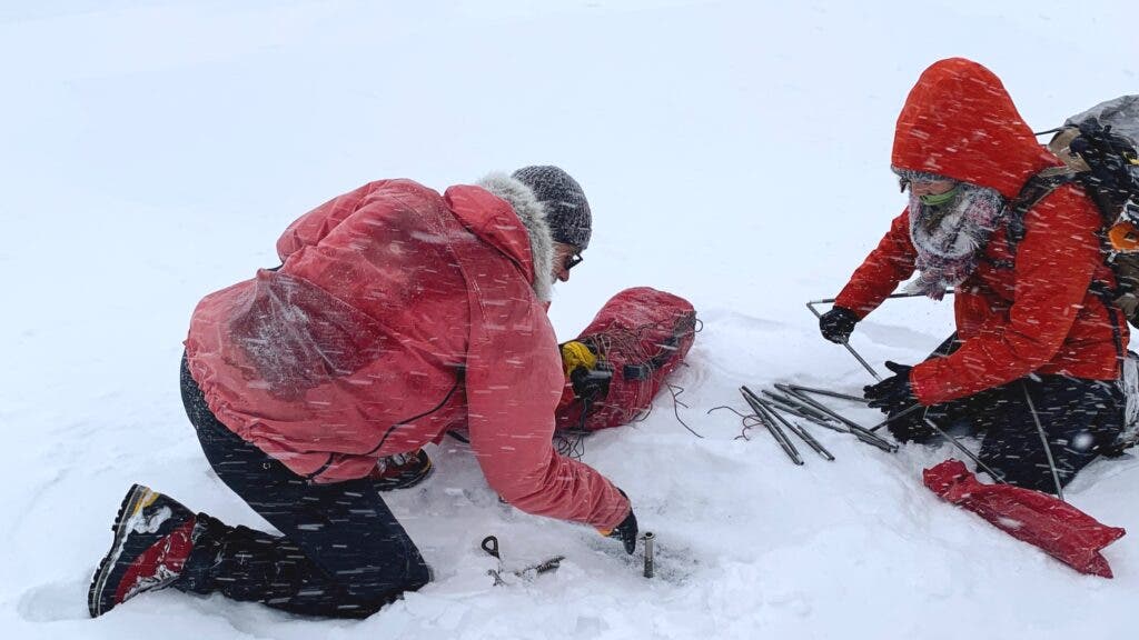 Two people wearing red jackets, on their hands and knees amid a snowstorm in Greenland, trying to set up their tent.