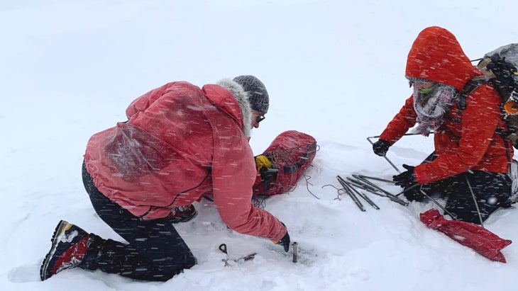 Two people wearing red jackets, on their hands and knees amid a snowstorm in Greenland, trying to set up their tent. 