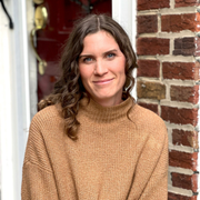 a headshot of a person with long brown wavy hair in a beige sweater looking at the camera