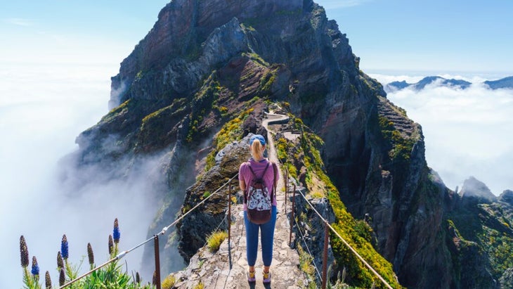 A woman hiking along a sideline trail to Pico Ruivo, Madeira. Clouds cover the valleys to either side.