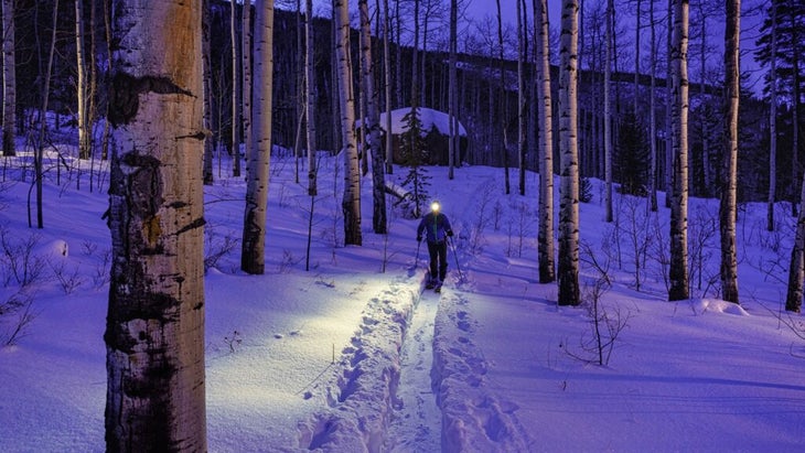 A backcountry skier follows a trail through a grove of aspens from a cabin at night. They are wearing a headlamp.