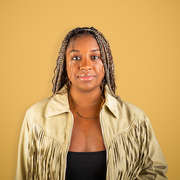 A woman with braids in a golden yellow shirt with a black shirt underneath.