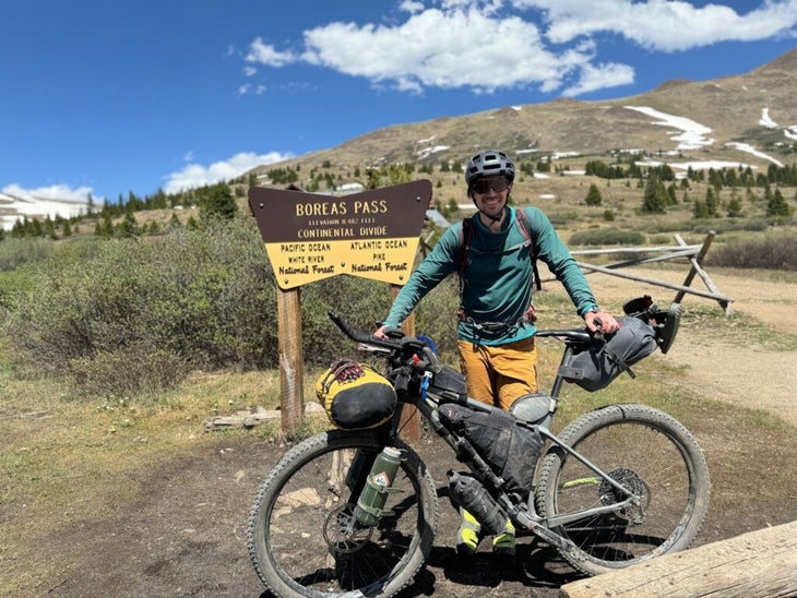 a man stands beside a bikepacking bike on the roadside on a sunny day