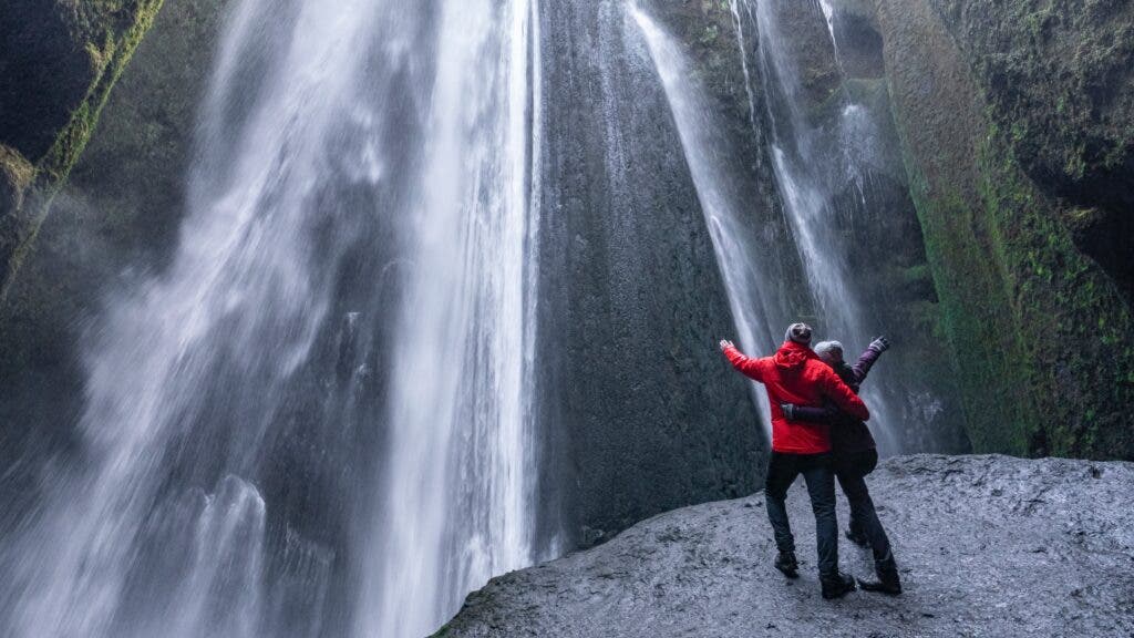 A couple embrace while on a rock at the base of a massive waterfall.