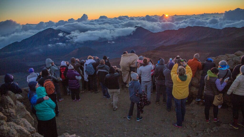 A crowd of people wearing jackets, some bundled in sleeping bags, watch the sunrise from atop Maui's Haleakala Crater.