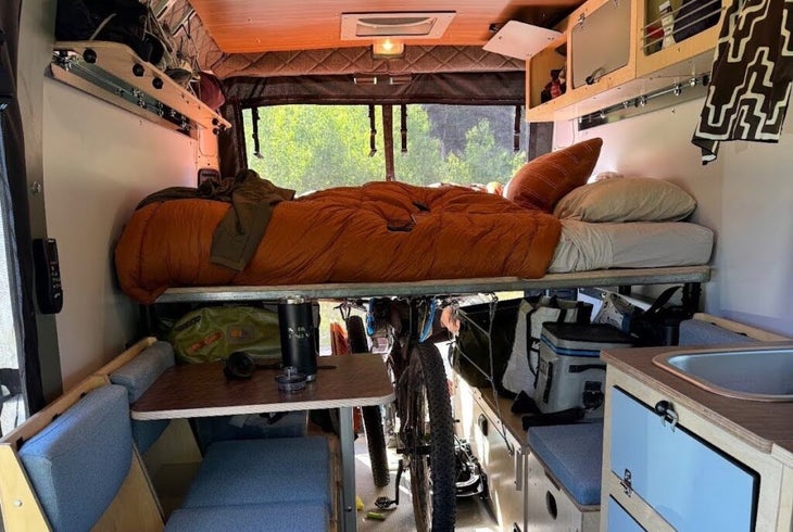 The interior of a camper van with blue cabinets and orange bedding during the daytime