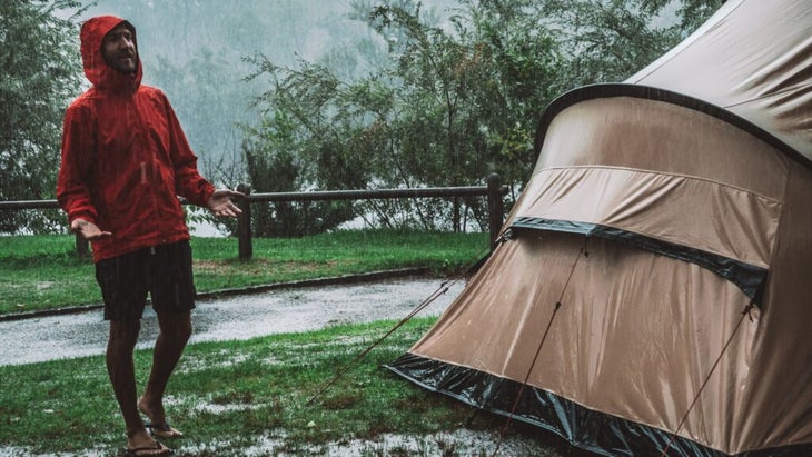 A man wearing a red rain jacket stands outside of his drenched tent in the pouring rain.